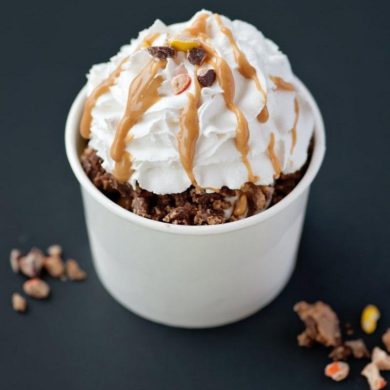 Des céréales marrons dans une tasse blanche, avec sur le dessus de la crème chantilly avec un liquide brun. Le tout sur une table noire