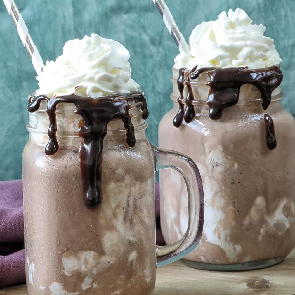 2 tasses remplies d’une boisson au chocolat et sur chacun il y a de la crème chantilly blanche et des pailles blanches et argentés. Le tout sur une table en bois clair